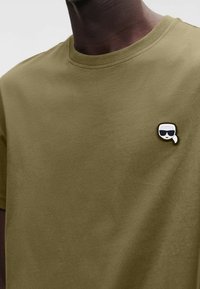 Olivgrünes Baumwoll-T-Shirt mit rundem Ausschnitt, das auf der linken Brustseite eine schwarz-weiße Anstecknadel mit einem Gesicht mit Sonnenbrille zeigt.