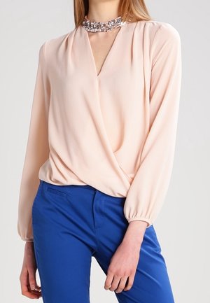 Mujer que lleva una blusa rosa pálido de manga larga con cierre cruzado en el frente y pantalones azules, complementada con un collar choker brillante.