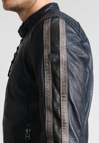 Námořnická kožená bunda s kontrastními černými pruhy a texturovaným světle šedým akcentem. Obsahuje zip a logo nášivku na rameni.