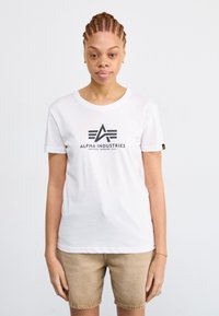 T-shirt bianco in cotone con scollo rotondo, maniche corte e logo nero "Alpha Industries" sul davanti. Indossato con pantaloni corti beige.