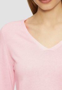 V-Ausschnitt Pullover aus weichem rosa Strick, mit glatter Textur und figurbetonter Silhouette. Dezente Nahtdetails an den Schultern.