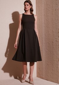 Czarna, bezrękawnikowa sukienka w kształcie litery A z kwadratowym dekoltem, plisowanym dołem i gładką fakturą. Noszona z beżowymi szpilkami z akcentami w postaci kokard.
