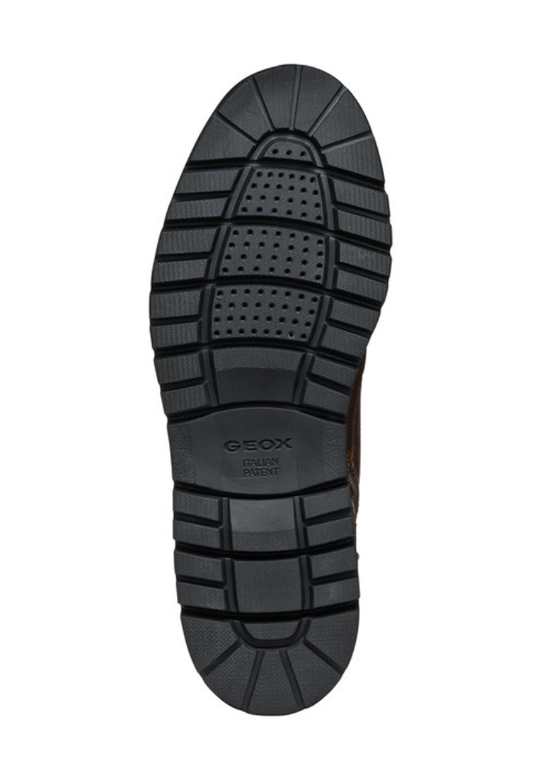 Semelle en caoutchouc noir avec un profilé texturé, comportant des rainures et des perforations pour la respirabilité. Logo de la marque embossé près du centre.