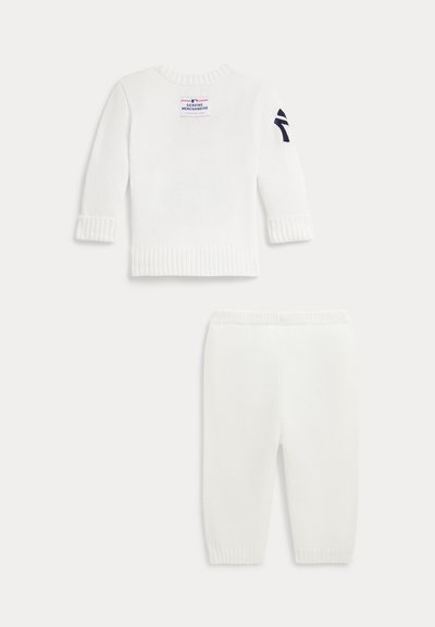 Conjunto de bebé blanco de punto con suéter de manga larga que presenta un pequeño parche en la espalda y un logo azul marino en la manga izquierda, acompañado de pantalones a juego.