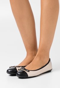 Ballerines avec un corps crème et un bout noir. Présentent un nœud avec de petits accents métalliques à l'avant. Texture lisse, design bas.