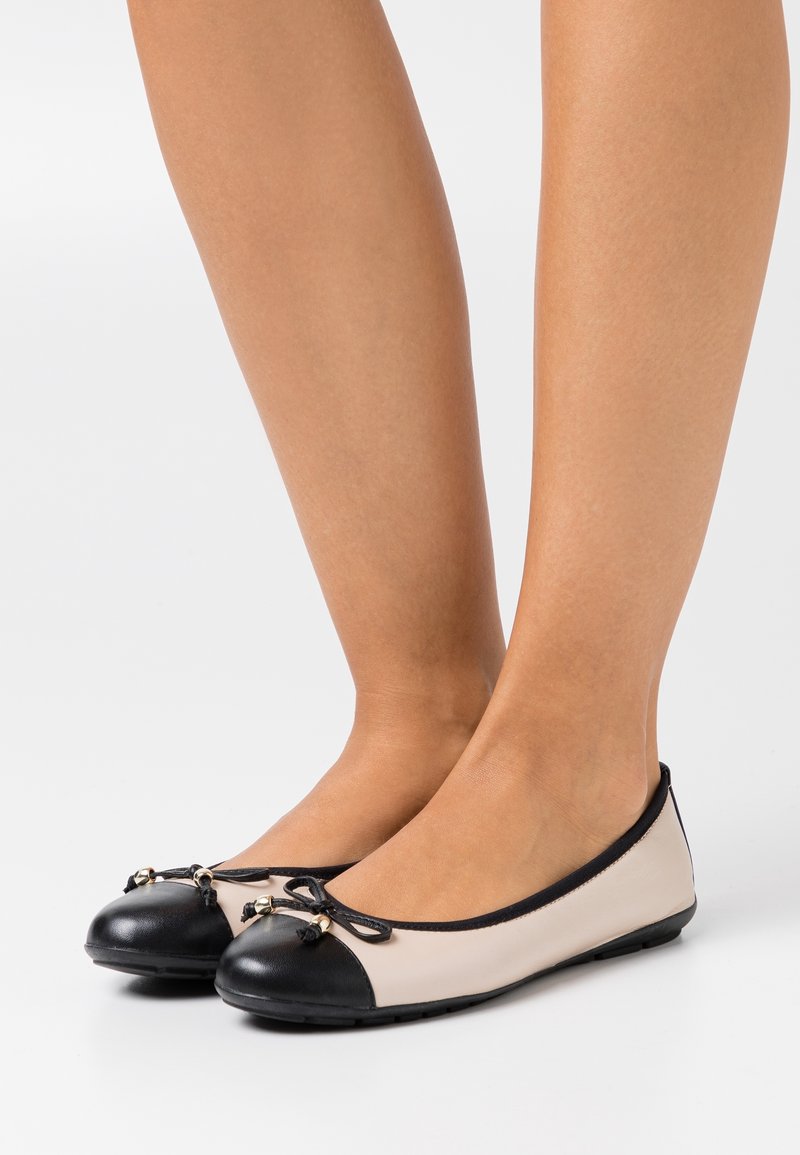 Ballerines avec un corps crème et un bout noir. Présentent un nœud avec de petits accents métalliques à l'avant. Texture lisse, design bas.