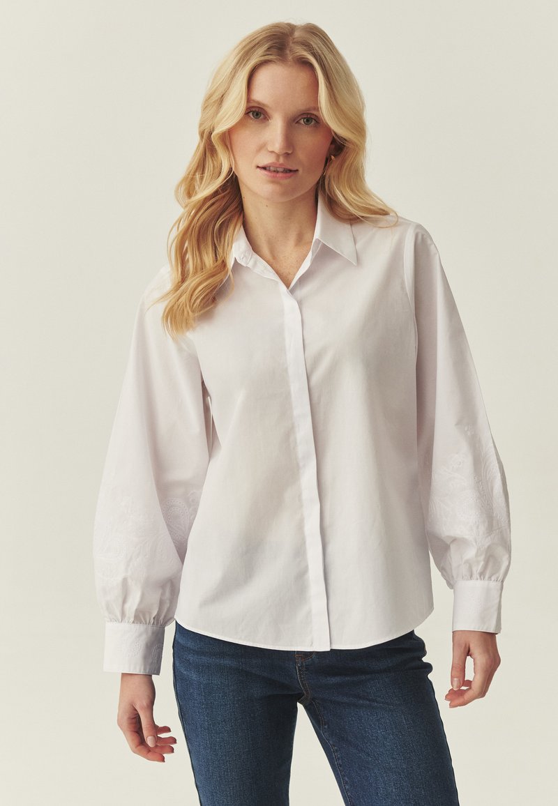 Chemise blanche à boutons avec manches longues bouffantes ornées de broderies sur les poignets. Le matériau semble lisse et léger.