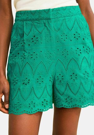 Pantaloni scurți verzi cu talie înaltă, cu decupaje florale brodate și tiv ondulat, purtați cu un top alb pe o persoană cu pielea închisă la culoare.