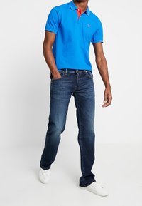 Camisa polo azul con acentos en el cuello rojos, combinada con jeans azul oscuro y zapatillas blancas. La camisa tiene un logo en el lado izquierdo del pecho.