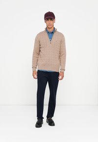 Jonge man in een bruine kabelgebreide trui over een blauw denim shirt, marineblauwe broek, zwarte instappers en een donker bordeauxrode pet, staand tegen een witte achtergrond.
