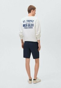 Vit sweatshirt med blå grafisk text på baksidan, tillsammans med marinblå shorts. Modellen bär ljusa skor med gröna accenter. En enkel design.