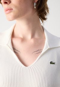 Weißes geripptes Strickoberteil mit V-Ausschnitt und umgeschlagenem Kragen, versehen mit einem grünen gestickten Logo auf der linken Seite. Haut mit sichtbaren Tattoos.