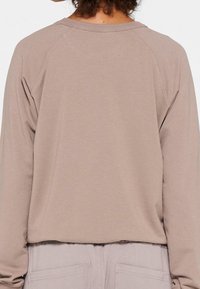 Sudadera de manga larga en color taupe claro, con cuello redondo, mangas raglán y una tela suave y texturada. Diseño cropped que termina por encima de la cintura.