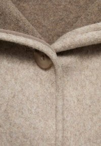 Beige vlna tkanina kabátu s měkkou texturou, s hnědým knoflíkem a kulatým límcem s kontrastním lemem.