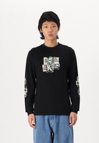 T-shirt noir à manches longues avec un design graphique sur le devant et les manches, présentant un texte coloré et un motif camouflage. Porté avec un jean bleu clair.