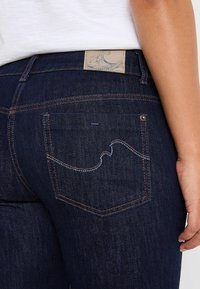 Jeans de mezclilla azul oscuro que presentan un diseño distintivo en el bolsillo trasero con costuras en azul claro y una etiqueta marrón con un gráfico de ola.