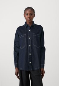 Mörk denimskjorta med krage, framslutning med knappar, två bröstfickor och kontrasterande vit sömnad. Långa ärmar med knäppta manschetter.