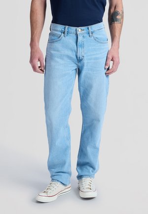 Homme portant un jean droit bleu clair, une chemise bleu marine, des baskets blanches et un tatouage noir sur son avant-bras droit.
