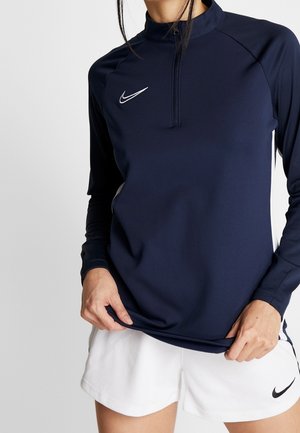 Personne portant un haut de sport bleu marine à manches longues avec une fermeture éclair devant et un short blanc, tous deux avec le logo Nike.