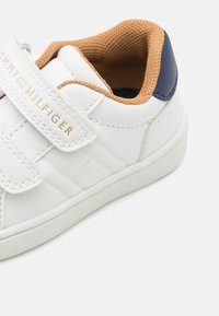 Białe skórzane sneakersy z zapięciem na rzep, granatowym akcentem na pięcie i brązowym, teksturowanym wnętrzem. Posiadają subtelną logo i przeszycia.