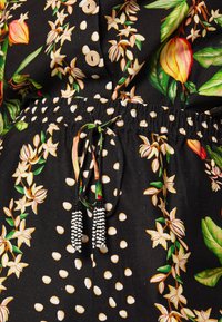 Tela negra floral con lunares blancos, con cintura ajustable mediante cordón y acentos de cuentas y botones de madera.