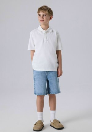 Junge steht mit den Händen hinter dem Rücken, trägt ein weißes Poloshirt, hellblaue Jeansshorts, weiße Socken und beige Slip-on-Schuhe.