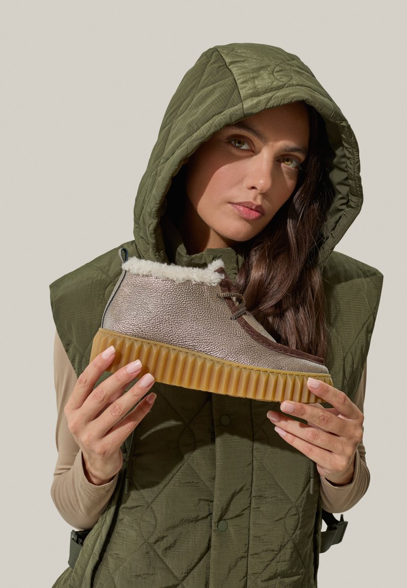 Brązowe i metalicznie srebrne botki za kostkę z teksturowanym cholewką, sztucznym futrem w środku oraz żebrowaną gumową podeszwą, trzymane przez osobę w oliwkowej kamizelce.