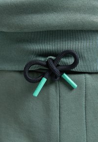 Zelená bavlněná tkanina s žebrovaným pasem má černou šňůrku uvázanou do mašle, doplněnou světle zelenými plastovými koncovkami.