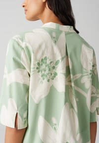 Light green blouse featuring large white floral patterns, short sleeves with side slits, and a casual collar design. Soft fabric texture.