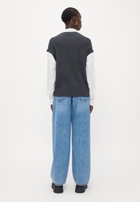 Grå strikket vest worn over hvit skjorte, kombinert med lyseblå vide denimjeans og svarte klumpete sko. Enkelt design og teksturert stoff.