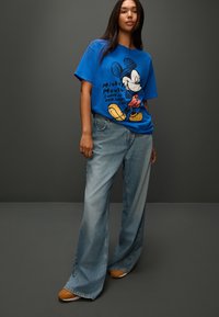 Blue cotton graphic t-shirt featuring Mickey Mouse design with text, paired with light-wash denim jeans and brown shoes.