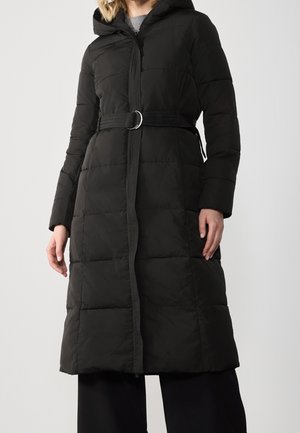 Femme portant un long manteau matelassé noir avec capuche et taille ceinturée, debout devant un fond clair uni.