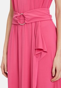 Rotes Kleid mit einem gerafften Gürtel, der mit einem silbernen Ring verziert ist. Weicher, fließender Stoff mit drapierten Details an der Seite und glatter Textur.