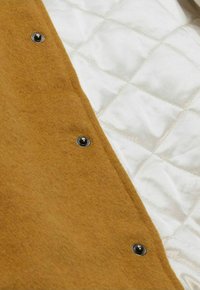 Brown wool fabric with a quilted cream lining, secured by black metallic snaps. Visible texture contrasts between materials.