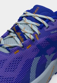 Chaussure de course violette avec dessus en mesh, rayures latérales texturées, lacets accentués et une semelle crème ornée d'un logo orange.