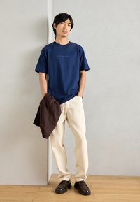 Ναυτικά μπλε βαμβακερό μπλουζάκι με λογότυπο, μπεζ παντελόνι, καφέ ελαφρύ μπουφάν κρεμασμένο στον αγκώνα και σκούρες καφέ loafers πάνω σε ξύλινο πάτωμα.