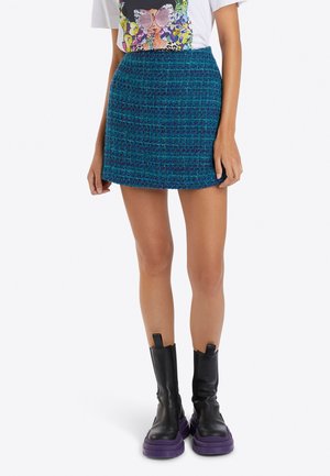 Kvinde iført blå tekstureret mininederdel, hvid T-shirt med grafik og sorte støvler til midt på læggen med tykke lilla såler, stående mod en hvid baggrund.