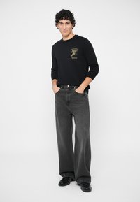 Νεαρός άνδρας με τα χέρια στις τσέπες, φορώντας μαύρη μπλούζα με γραφικά, φαρδιά γκρι τζιν και μαύρα παπούτσια, σε απλή λευκή φόντο.