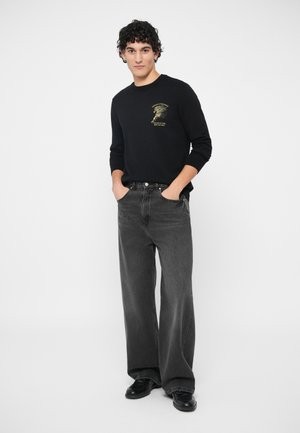 Noor mees seisab käed taskus, seljas must graafikaga sviiter, laiad hallid teksad ja mustad kingad, taustal valge sein.