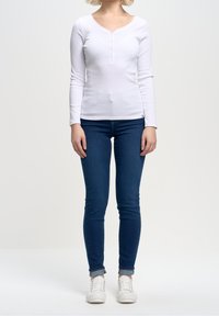 Long-sleeved white ribbed top with a V-neck and five-button placket, paired with blue skinny jeans and white sneakers.
