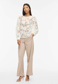 Femme portant un chemisier blanc transparent à fleurs avec des manches bouffantes, un pantalon beige à jambes larges et des talons ouverts argentés, debout avec une main dans la poche.