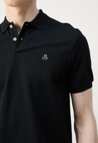 Polo noir à manches courtes porté par une personne, présentant un logo brodé blanc "M O P" sur la poitrine gauche et deux boutons de couleur crème sur la patte de boutonnage du col.