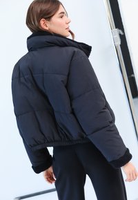 Giacca imbottita corta nera con collo e polsini pelosi e strutturati. Caratterizzata da un design trapuntato e una vita elastica per una vestibilità aderente.