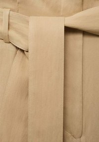 Beige tkanina s hladkým povrchem obsahuje uvázaný pásek. Design zahrnuje čisté linie a jemné šití, což zdůrazňuje jednoduchost.