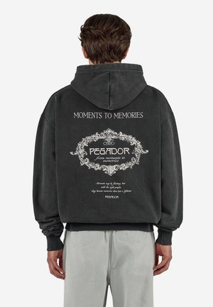 Mężczyzna noszący czarną bluzę z kapturem z białym dekoracyjnym napisem na plecach: "Moments to Memories" oraz "PEGADOR from moments to memories."