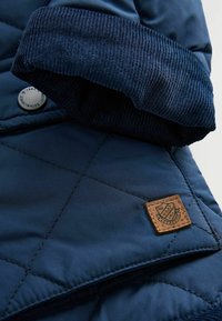 Mørkeblå quilted jakke med en kordfløjlsmanschet, der har en firkantet lædermærke og metaltrykknap, som fremhæver en glat, skinnende stofstruktur.