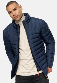 Navy quilted jacket with zip closure and high collar, featuring a beige sweater underneath and orange pull-tabs on zippers.