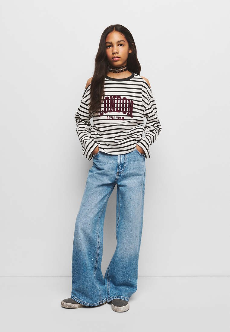 Jeune personne debout, les mains dans les poches, portant un top à épaules dénudées rayé avec le texte "London", jean à jambes larges et chaussures grises sur fond blanc.