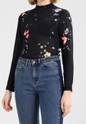 Kobieta ubrana w prześwitującą czarną bluzkę z długim rękawem z kwiatowymi wzorami oraz wysokim stanem niebieskich dżinsów na jednolitym tle.