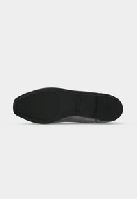 Semelle d'une chaussure formelle noire avec des zones de préhension texturées au talon et à l’orteil, présentée sur un fond gris clair uni.